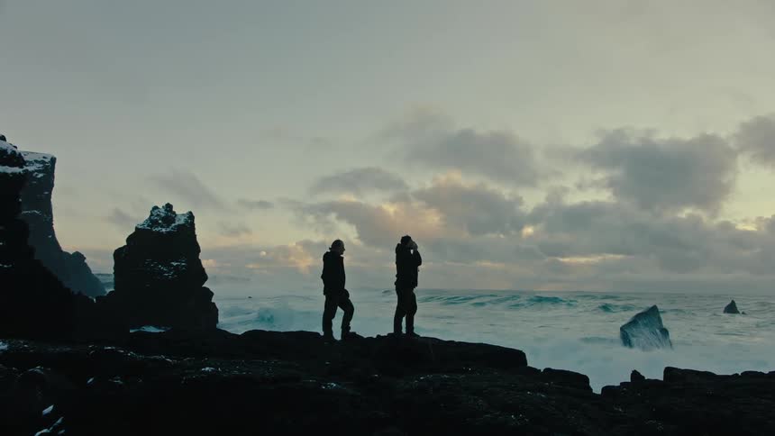 Two men are standing along the coast taking pictures as rough water waves crash into snow covered rocks along the shoreline