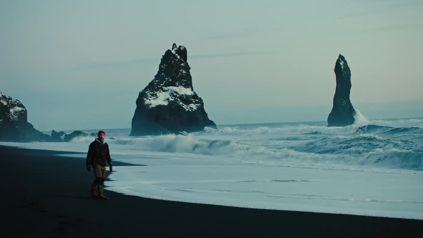 Man walking on black sand next to the sea in a place with snowcovered rocks
