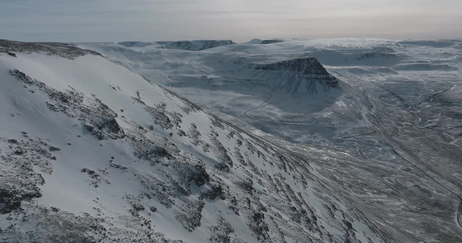 Snow covers the mountainside as a road runs through the river valley under a cloudy sky