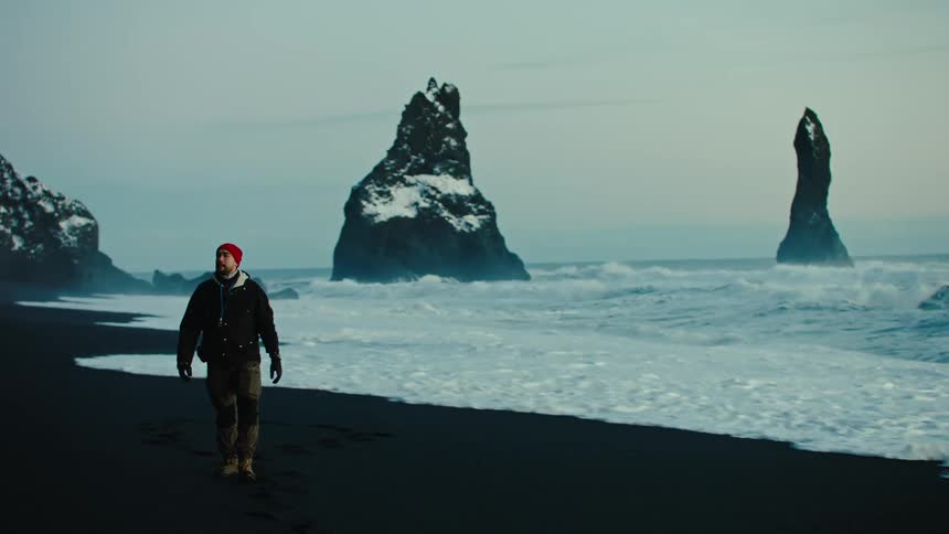 A man wearing a red hat walks on the beach at dusk