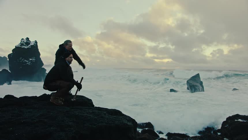 Two men on a rock looking at and discussing the stormy sea in front of them