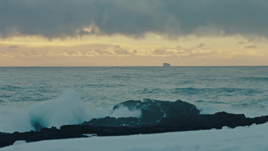 Dense clouds fill amber sky silhouetting iceberg a long distance from waves crashing against snowy coast