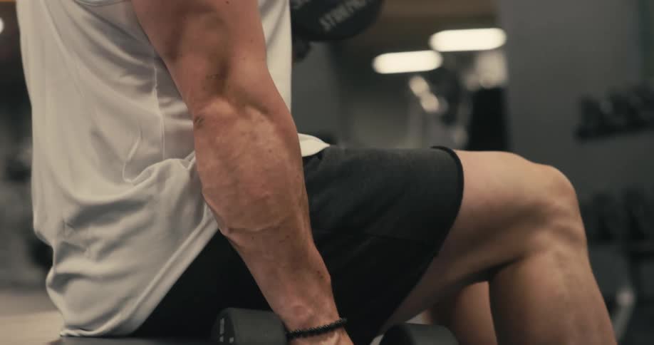 A man in a tank top sitting on a gym bench and performing weightlifting with dumbbells