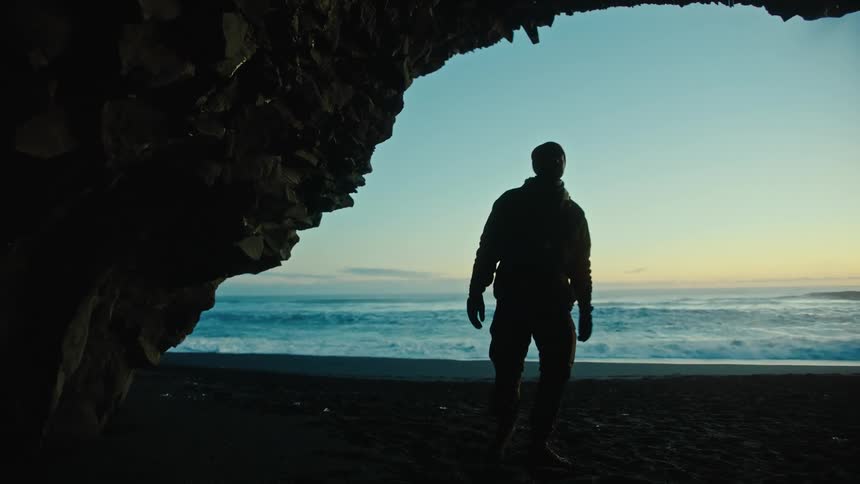 A man walking on the beach looks inside a cave as waves roll into shore behind him