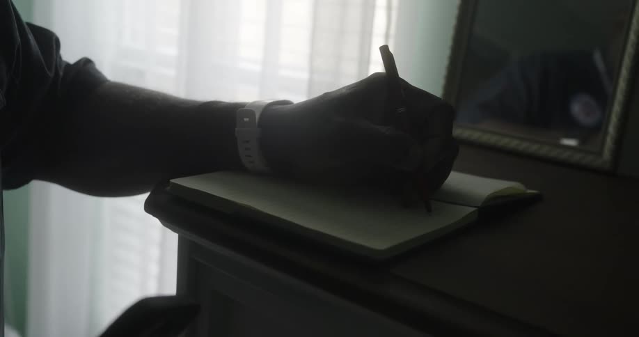 A person using a pen to write on a notebook on their dresser in the room as light comes through the window