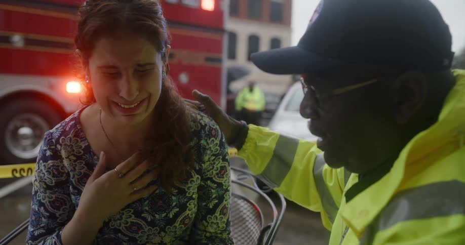 An emergency worker is comforting an upset woman on the side of a road with a police car with flashing lights and an ambulance in the distance