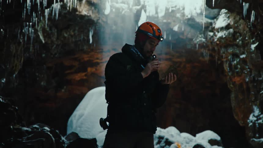 A man wearing a helmet examining a rock with a magnifier in an icy cave