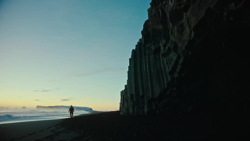 A man walking on a beach in the morning near a tall rock cliff as waves crash against the beach