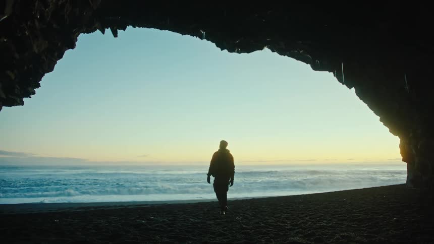 A man walking on a beach under a cove as waves crash against the coast behind him