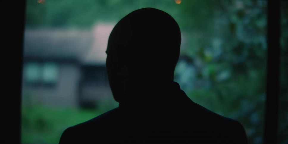 Silhouette of a bald man standing up outdoors next to a lawn with trees in front of a house