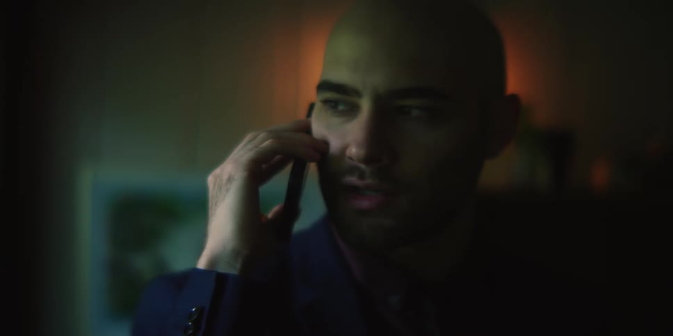 A man is standing in a dimly lit room turning his head as he talks on his phone