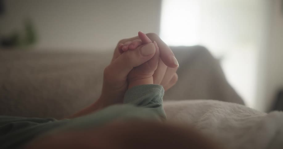 Close up of a womans hand holding her infant babies hand as they lounge together