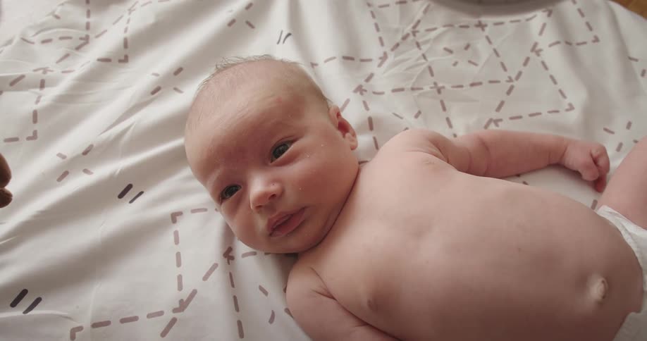 A infant baby layingon their back on a sheet with only a diaper on while looking around and stretching arms out