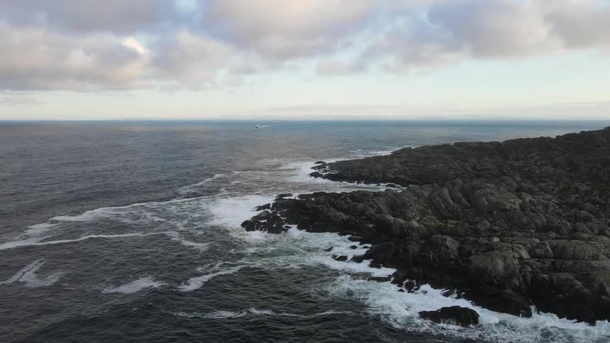 Aerial of ocean waves crashing against a rocky coast in the middle of the day with clouds covering the sky