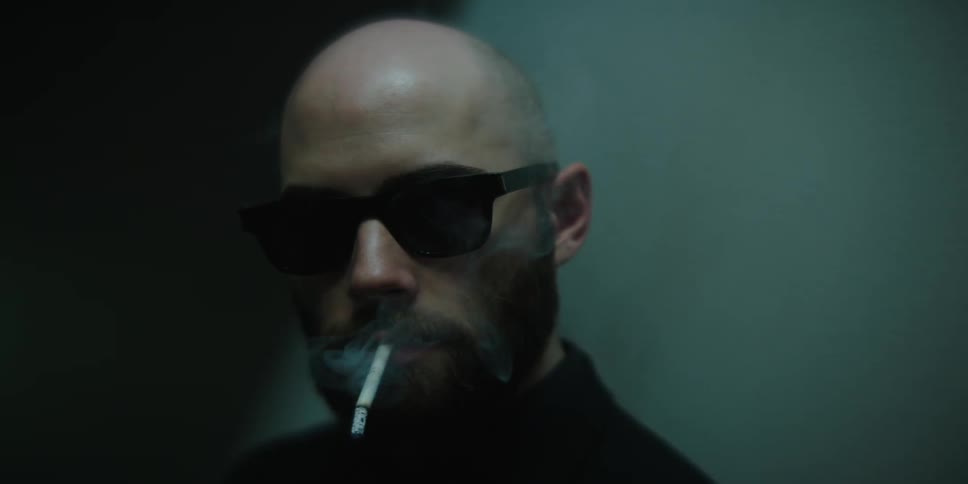 A man is wearing sunglass and smoking a cigarette in a room near the wall