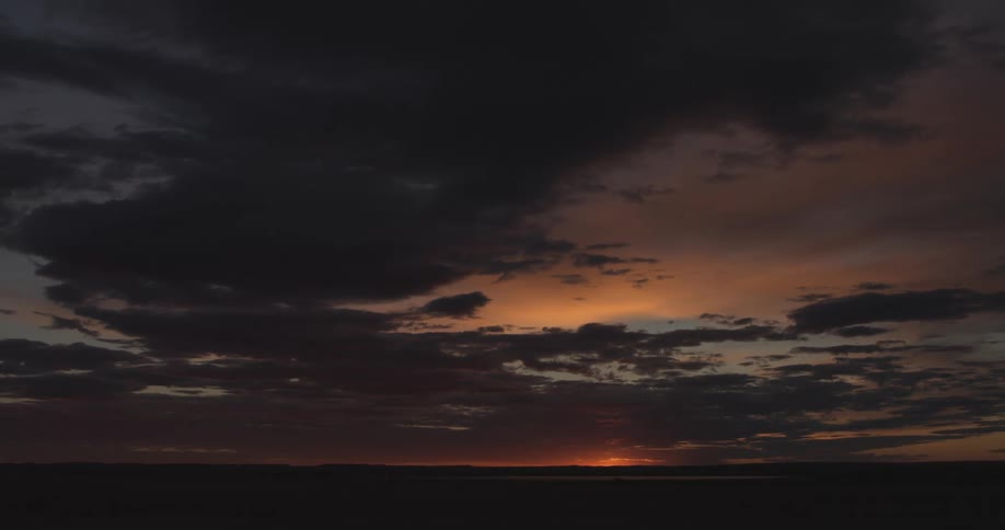 The sun rising on the horizon over a wide open landscape with dark clouds in the orange sky