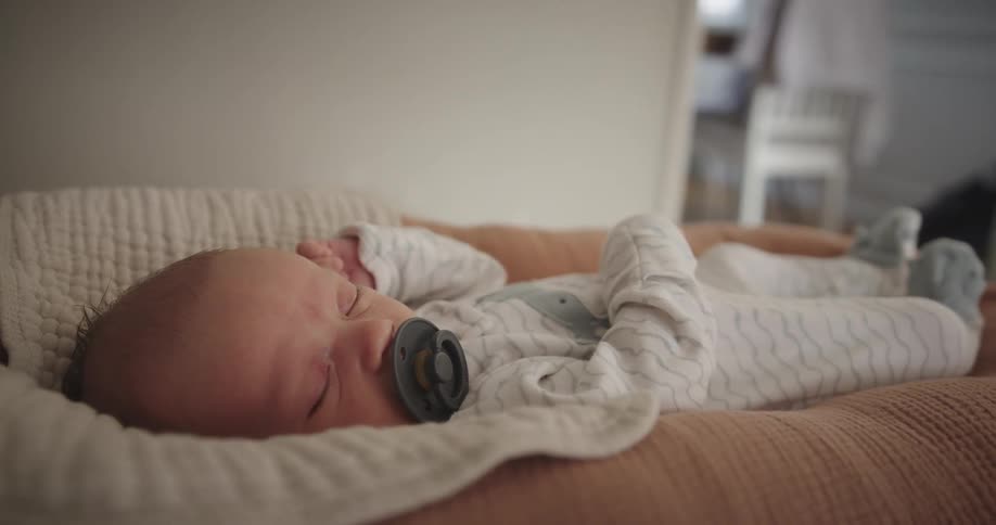 A infant baby with a pacifier in its mouth sleeping in pajamas on a bed