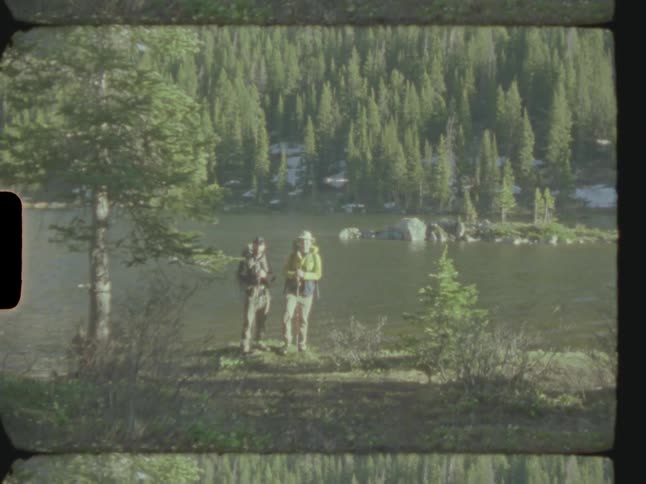 Two male hikers pose together near water across from conifer forest