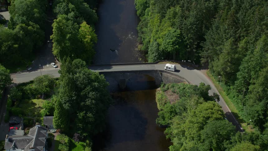 Vehicles drive along a bridge over a river flanked by dense trees and sprawling homes