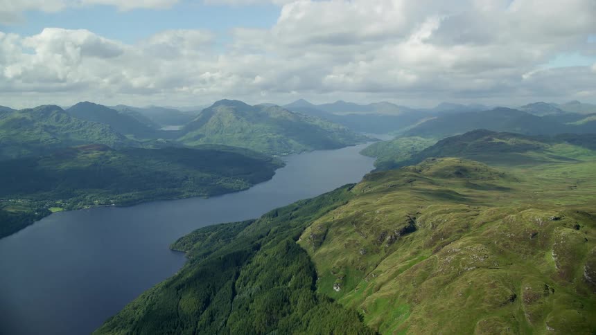 Drone view of a river flowing through a grassy mountain range with cumulus clouds covering the sunny sky