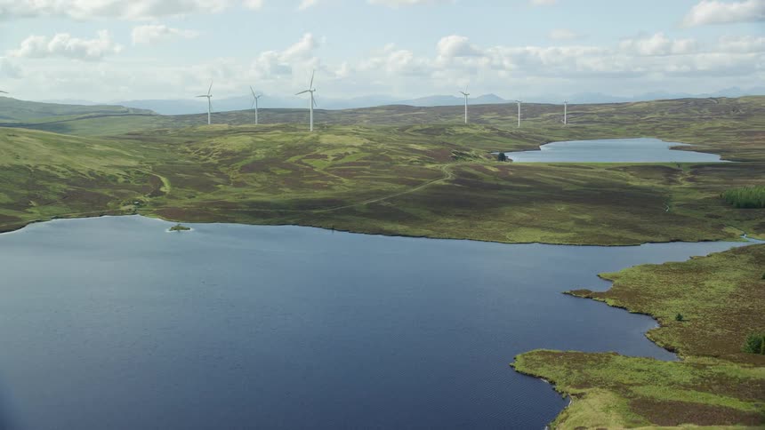 Wind turbines stand on hills near ponds of water with mountains in the distance