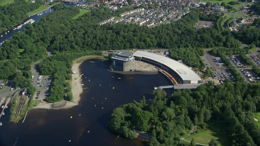 Aerial of a lake meeting land where an architectural building sits near a road and trees