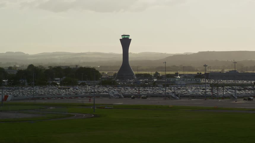 Aerial of a parking lot full of cars near an airport tower during the day with mountains on the horizon