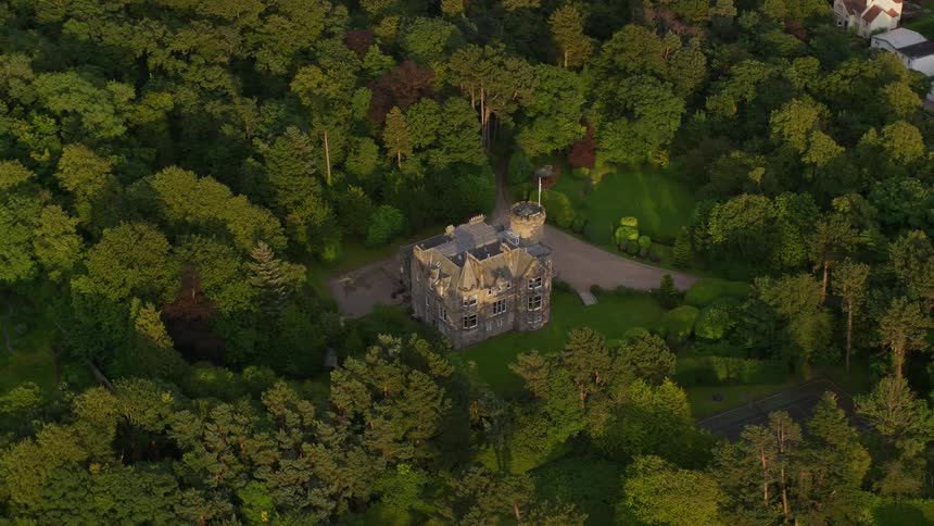 Aerial of a mansion built among a dense forest with tall trees as the sun shines on the house