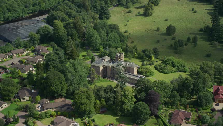 Aerial of a large manor surrounded by trees and other suburban households on a bright sunny day