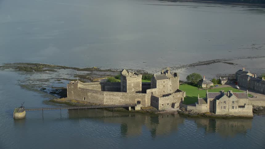 Aerial of a stone manor on the waters edge with tall stone walls surrounding it and a lush green lawn