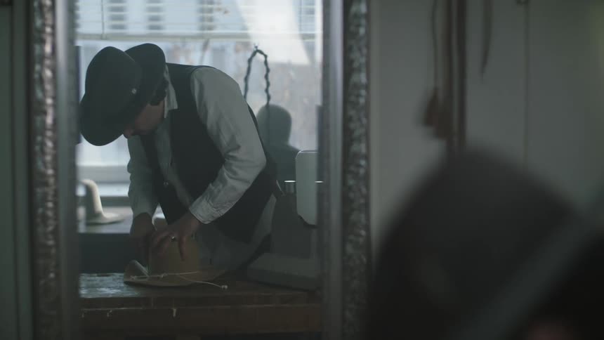Mirror reflects hatter working on hat in workshop