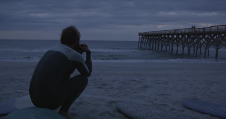 Man sits on his surfboard on the beach on a cloudy night
