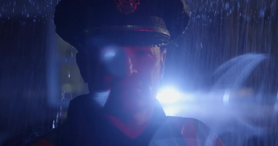 Policeman looking straightforward in the rain at night while the lights of his car flash behind him