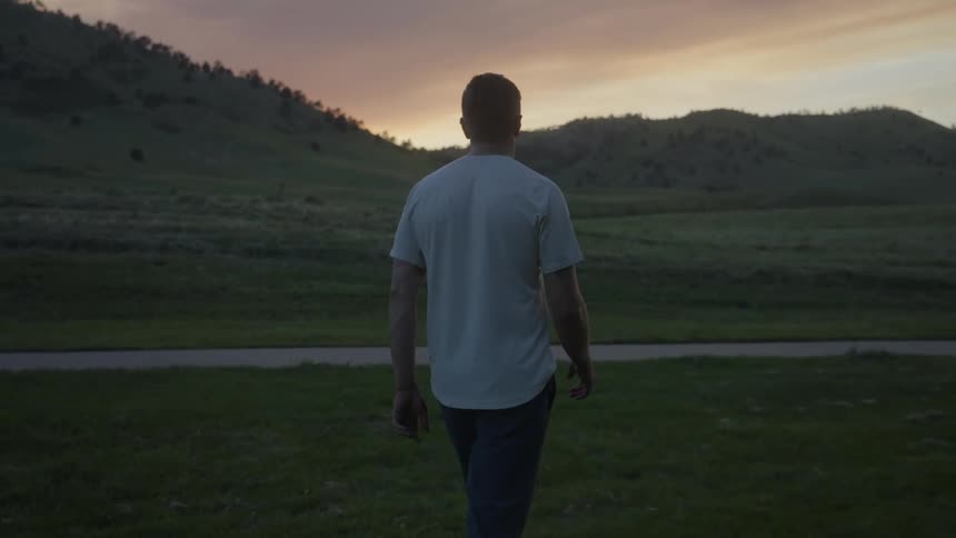 Man walks across field toward road and hills under a red tinted sky