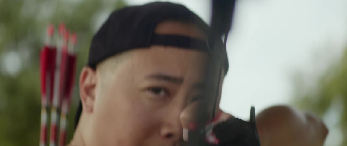 A man wearing a baseball cap raises a bow and aims an arrow
