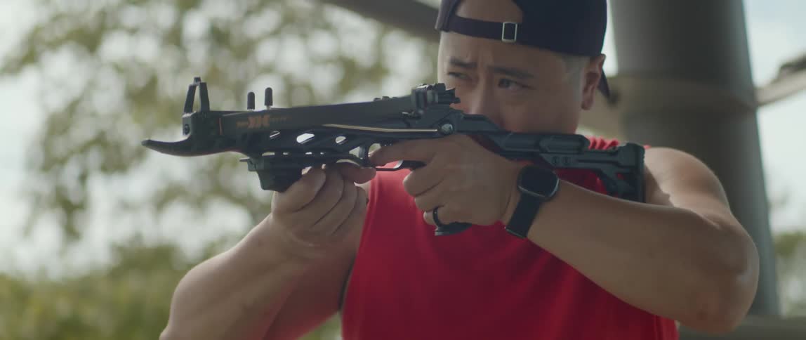 An asian man in a baseball cap puts an arrow in a crossbow and takes aim with it
