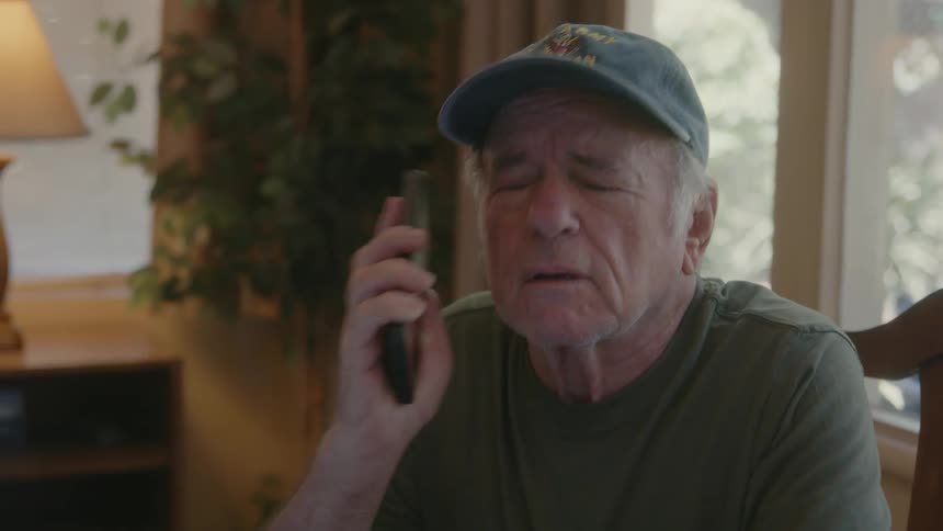 An elderly man is sitting in a chair in his living room talking on the phone as he pulls the phone away from his ear and rolls his eyes