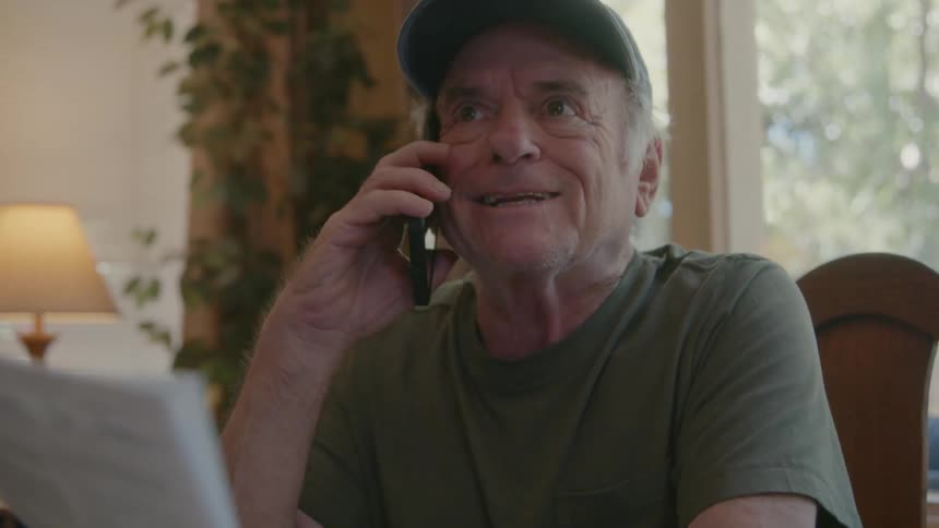 An elderly man talking with a cell phone to his ear while looking at a paper in his hands