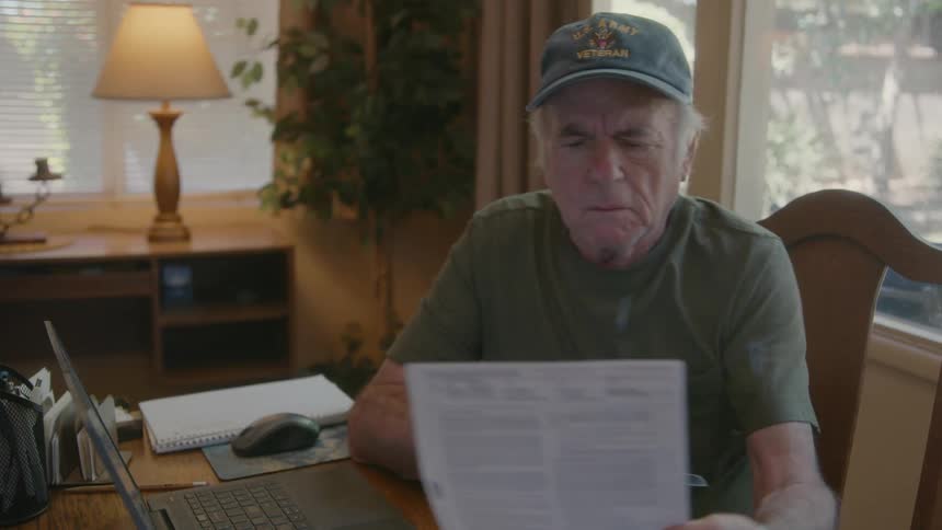An elderly person sitting down picking up their glasses to read a brochure