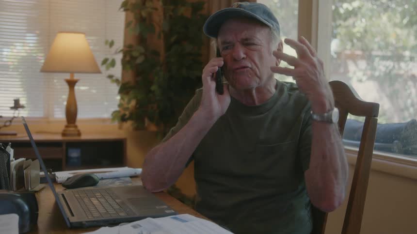 Man talking angrily on phone at midday while sitting on a chair indoor with a laptop opened