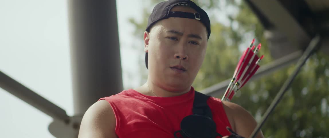 An asian man with a baseball cap pulls a bowstring to shoot an arrow
