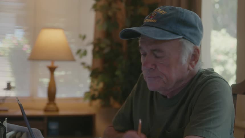 An elderly person sitting down and putting on their glasses to read a document