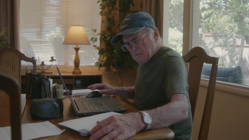 An elderly man in a baseball cap is sitting and typing on a laptop while going through some papers