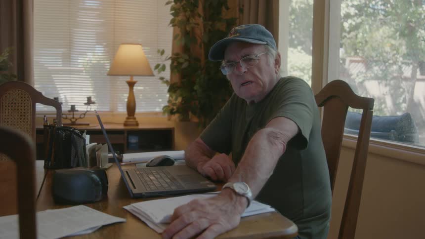 An elderly man sitting in front of a computer and looking at a paper