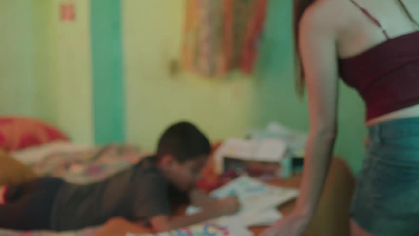 A woman walks up to her son who is lying on the bed drawing and talks to him