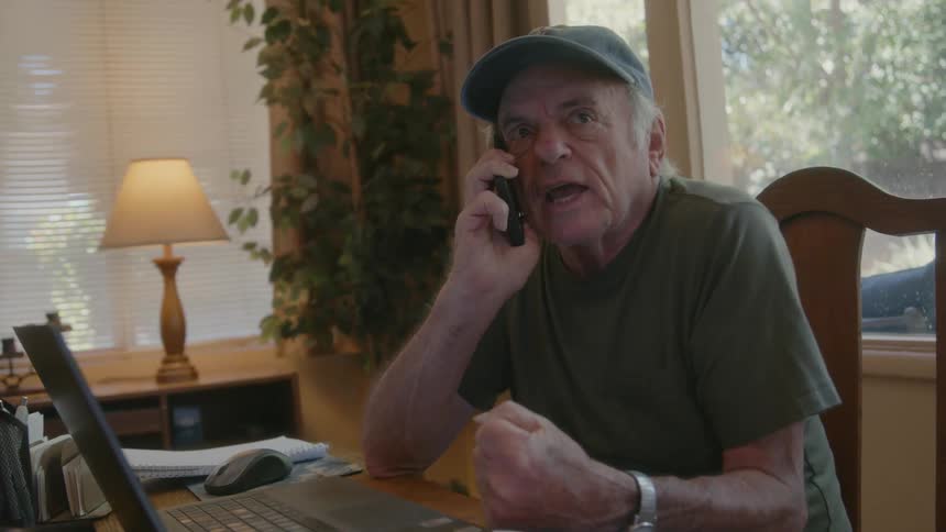Elderly man sits in home at table scattered with papers and talks aggressively on phone