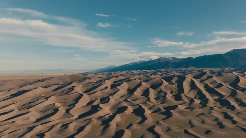 The sun shines upon ripples in the sand of the dunes with mountains in the distance under a cloudy sky
