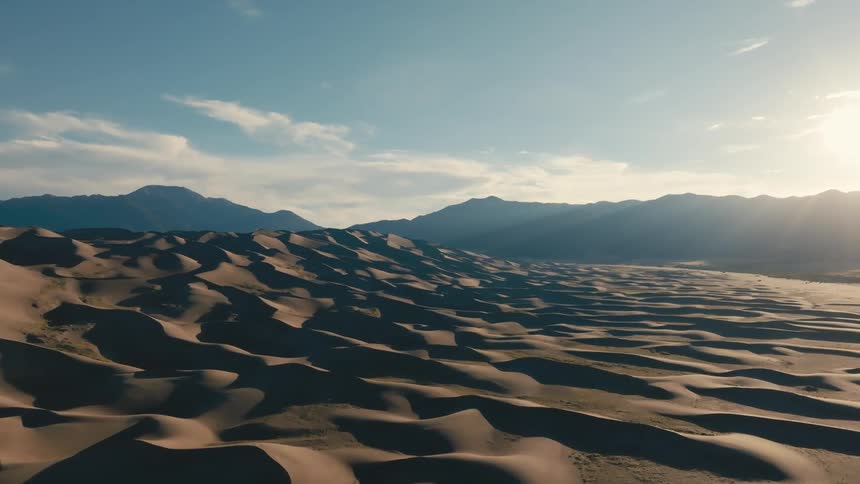 Aerial of dune covered landscapes with mountains on the horizon with the sun bright in the sky