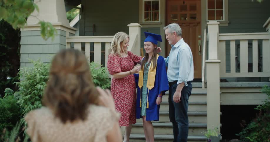 Woman uses smartphone to take a picture of parents posing with high school graduate in front of home