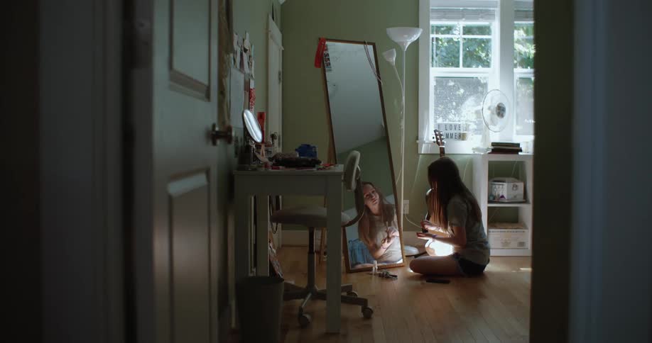 Teenage girl sits on floor at base of full length mirror and puts on makeup
